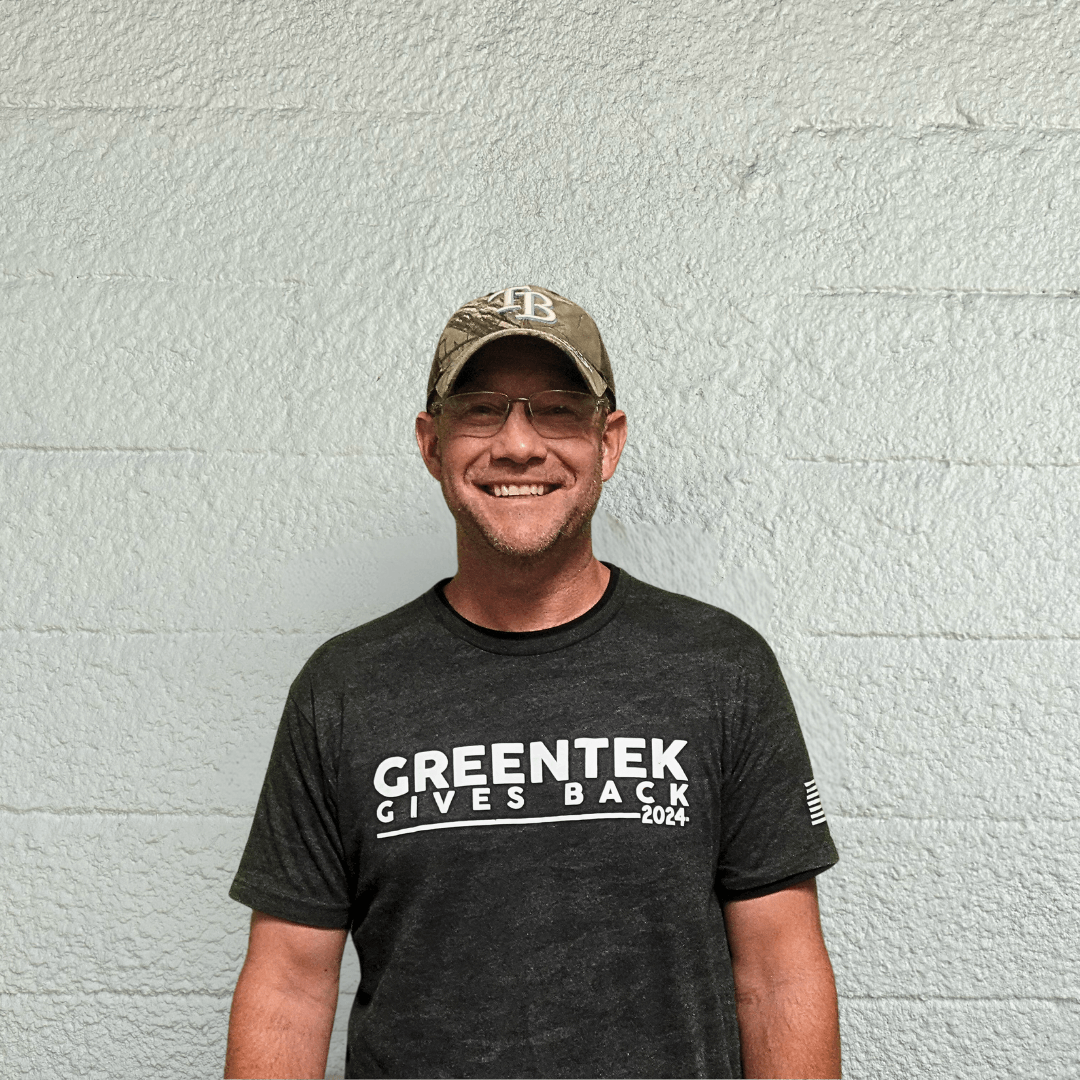 A smiling man wearing glasses, a gray cap, and a dark T-shirt that says "GREENTEK GIVES BACK 2024" stands in front of a light gray textured wall.