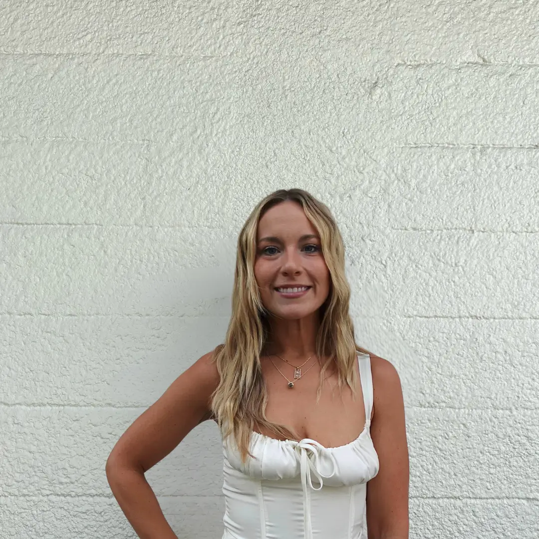 A woman with long, wavy blonde hair wearing a sleeveless white dress stands smiling in front of a textured white wall. One arm rests on her hip.