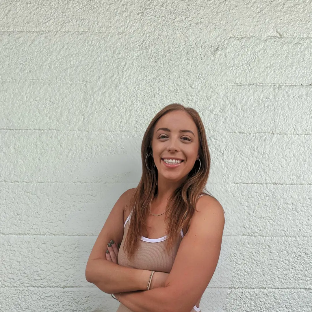 A woman with long brown hair smiles and stands with arms crossed in front of a textured white wall. She is wearing a light-colored tank top and hoop earrings.