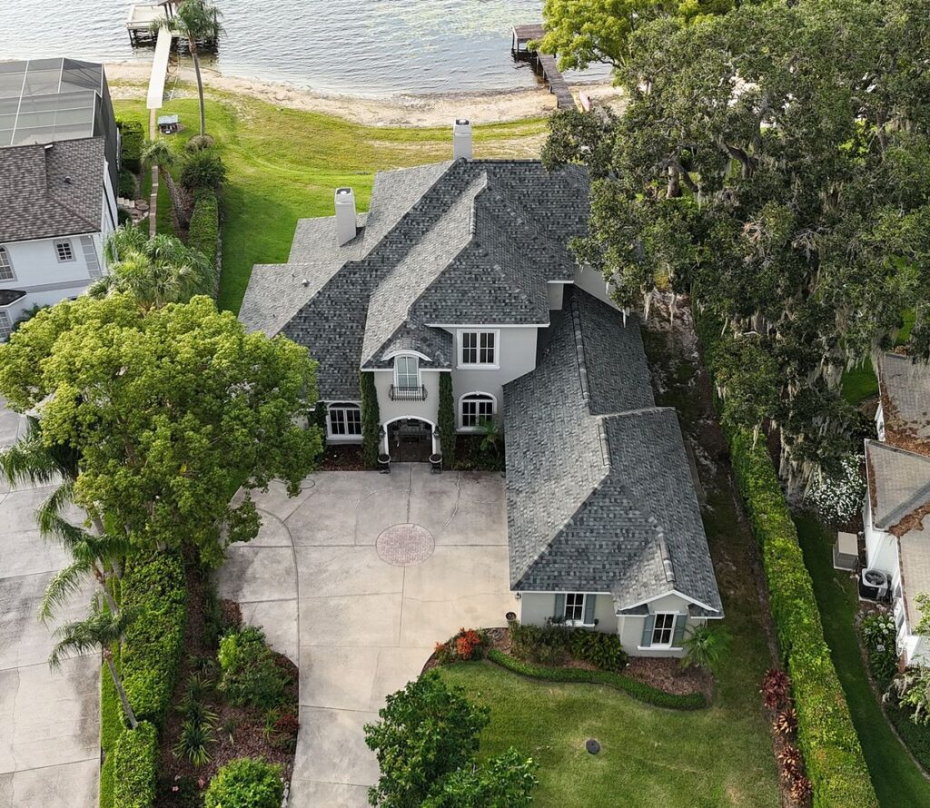 Aerial view of a two-story house with gray roof and light exterior, surrounded by trees and greenery, with a curved driveway, near a lake with a dock in the background.