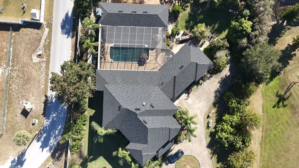 Aerial view of a large house with dark roofing, a screened-in backyard pool, curved driveway, and surrounding green trees and lawn. A street borders the property on the left side.