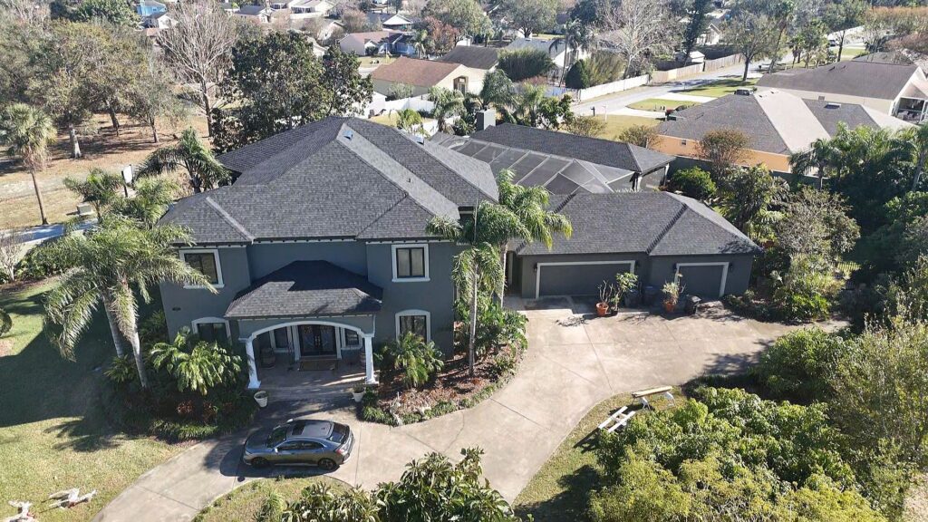 A large two-story house with a dark gray roof, surrounded by palm trees and greenery, with a curved driveway and a car parked in front. The home features multiple garages and is located in a suburban neighborhood.