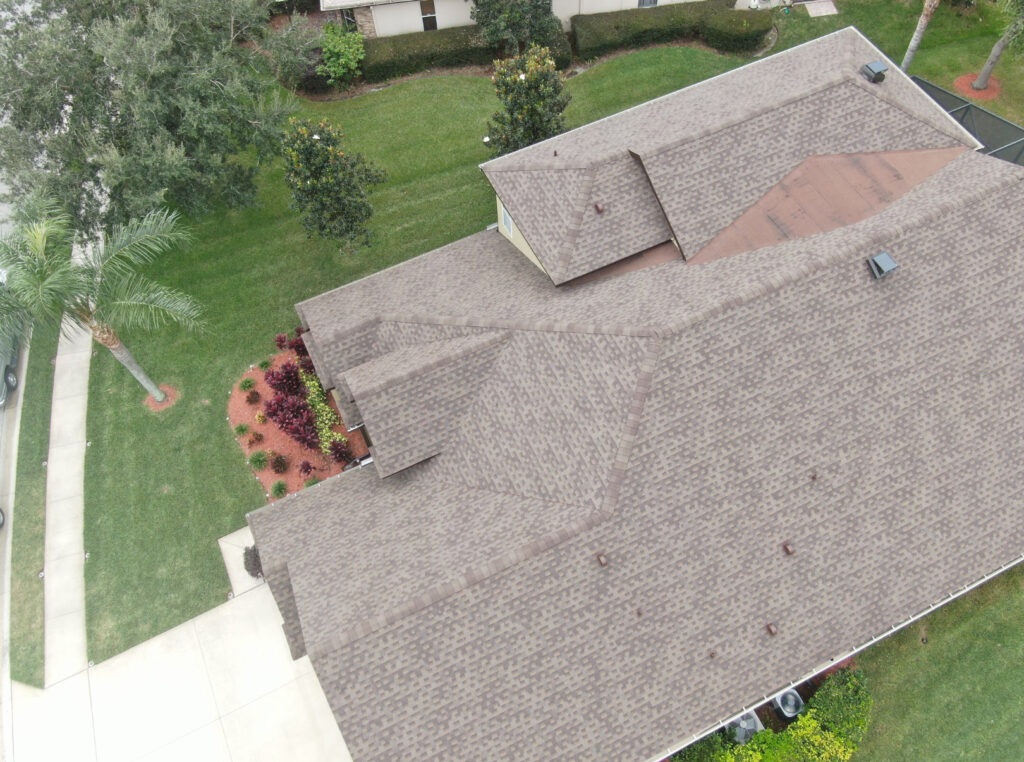 Aerial view of a house with a brown shingle roof, surrounded by green grass, trees, and landscaped shrubs near a driveway.