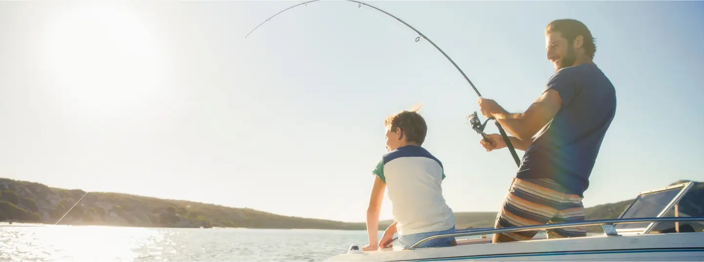 A man and a boy are fishing from a boat on a sunny day, with water and hills in the background. The man is holding a fishing rod while the boy sits on the edge of the boat, looking at the water.