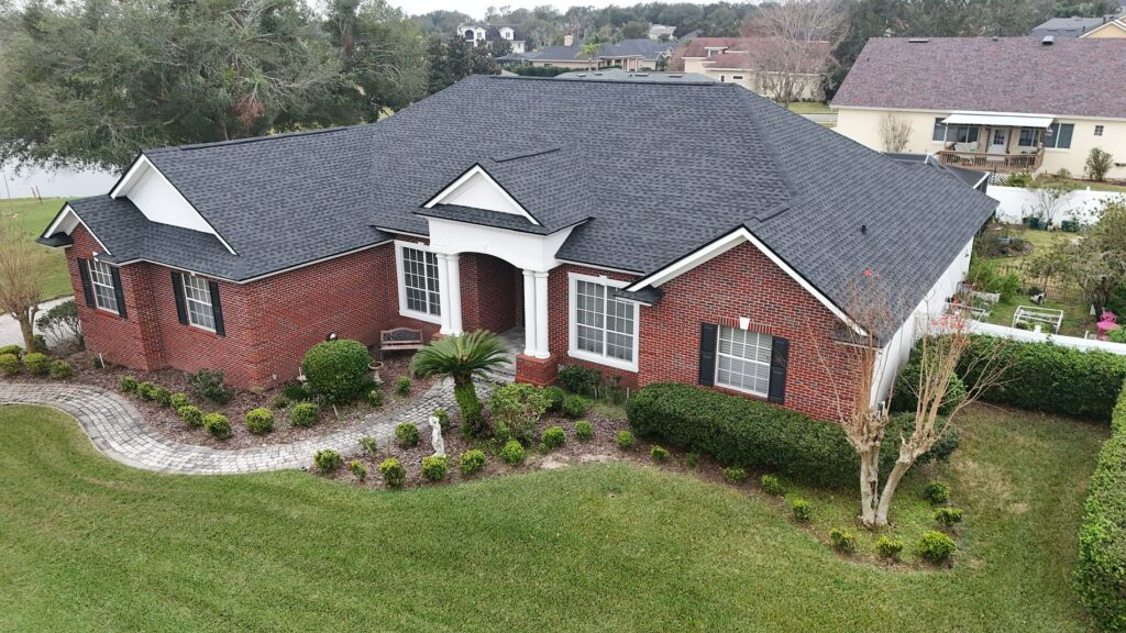 Aerial view of a single-story red brick house with black roof shingles, white trim, and manicured landscaping, including shrubs, a palm, and a curved stone walkway, set in a suburban neighborhood.