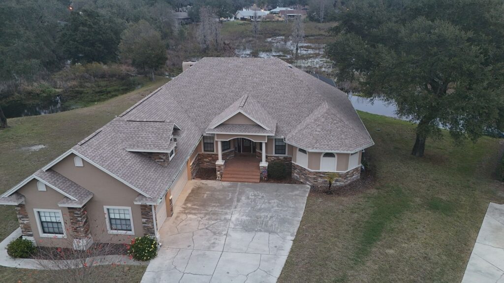 Aerial view of a single-story beige house with a large, sloped roof, a curved driveway, and landscaped front yard, surrounded by trees and near a small body of water in the background.