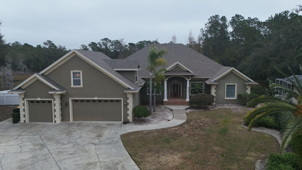 A large, two-story suburban house with a three-car garage, gray exterior, and a shingled roof. The front has columns, a curved driveway, some bushes, and palm trees, with trees in the background.
