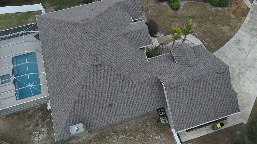 Aerial view of a house with a complex, gray shingled roof, a curved driveway, a small yard, and a screened swimming pool area on the left side.