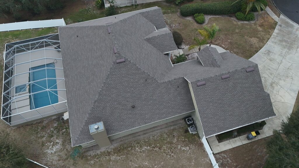 Aerial view of a house with a gray shingle roof, screened-in backyard pool, concrete driveway, and some surrounding trees and landscaping.