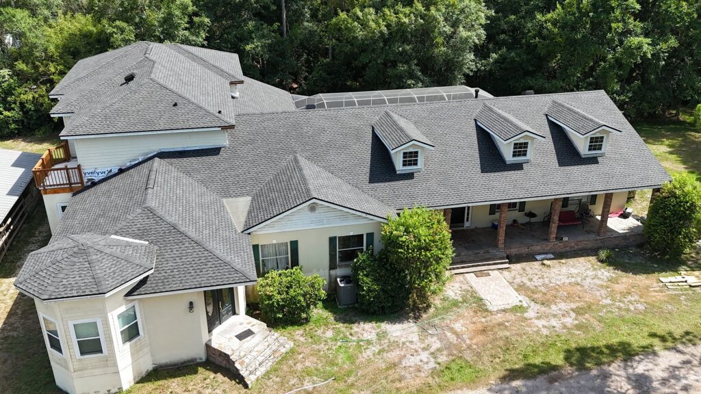 Aerial view of a large house with multiple gabled roofs, dormer windows, a screened-in patio, and a front porch with columns, surrounded by trees and a partially grassy yard.