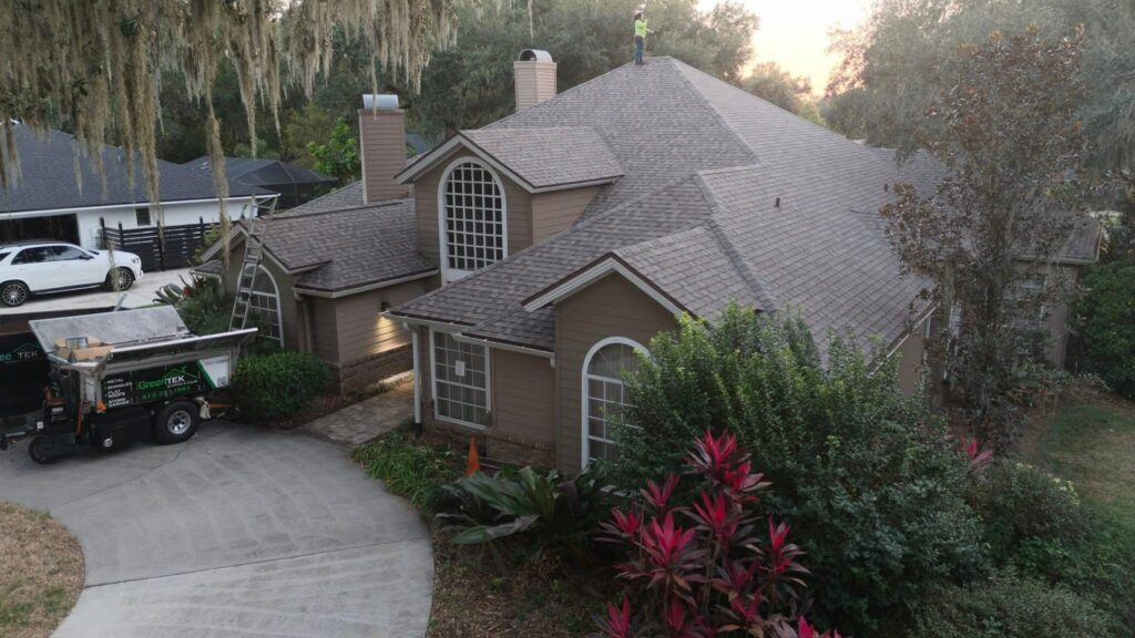 A person stands on the roof of a large, tan house surrounded by trees and plants, with a driveway curving in front and a truck labeled "GreenTek" parked nearby.