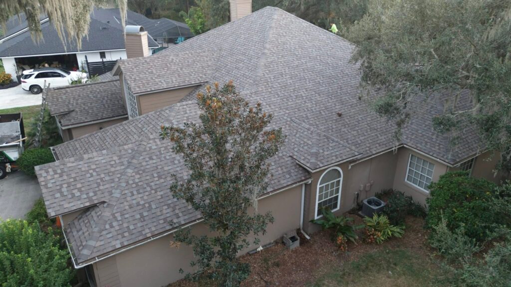Aerial view of a house with a gray shingle roof, beige exterior walls, large windows, and surrounding trees and shrubs. A white car is parked in a driveway next door.