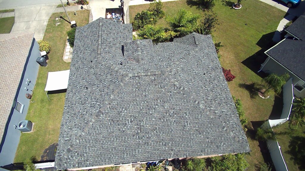 Aerial view of a house with a dark gray shingle roof, surrounded by a green lawn, trees, and neighboring homes in a suburban neighborhood.