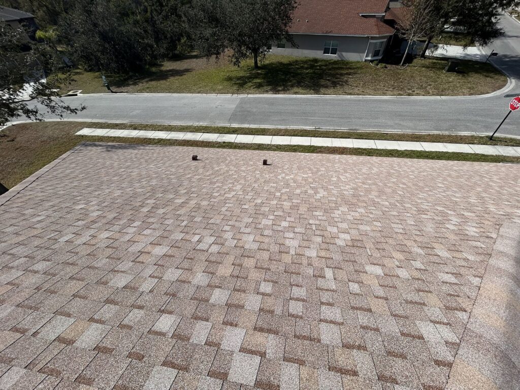 A view from a rooftop covered with brown asphalt shingles, overlooking a street, sidewalk, grass, and a house in the background. Two roof vents are visible near the center of the roof.