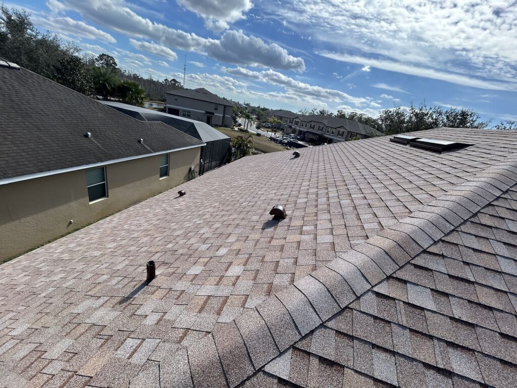 View of a residential neighborhood from a sloped rooftop with brown shingles, pipes, and a skylight under a partly cloudy blue sky. Neighboring houses and trees are visible in the background.