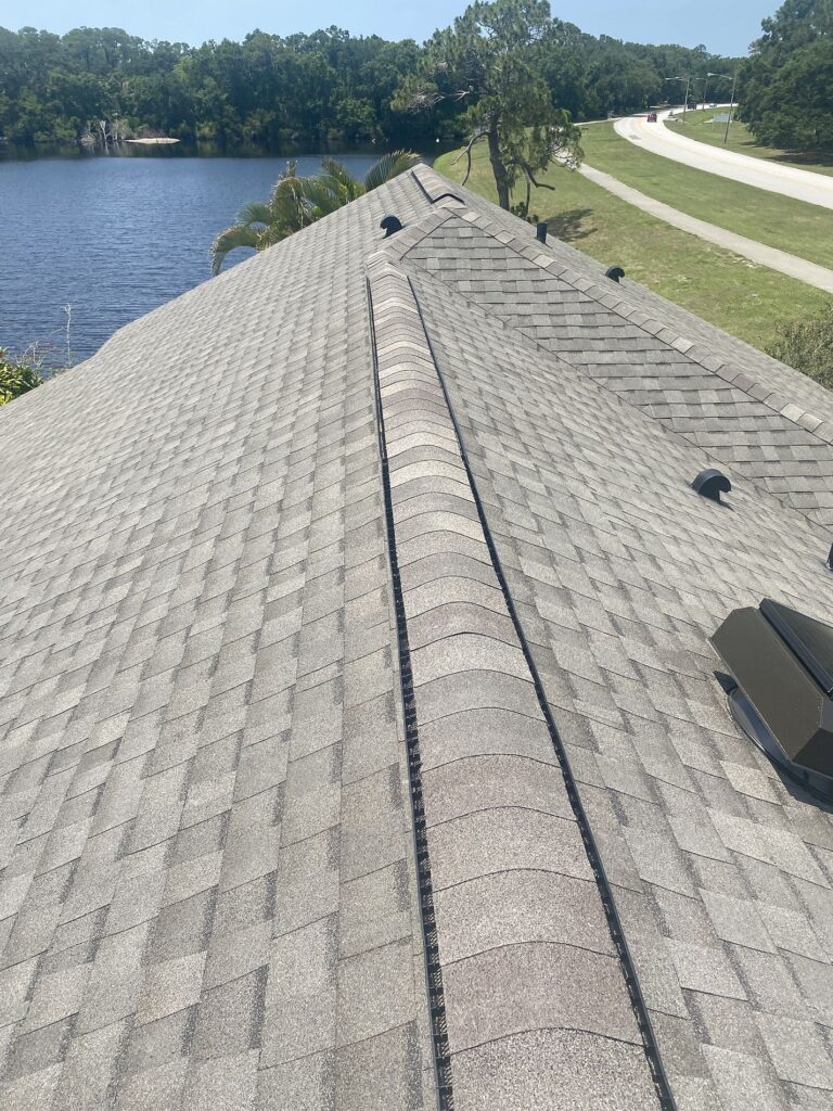 View from the peak of a gray shingled roof with vent caps, overlooking a lake, trees, and a road in the background on a sunny day.