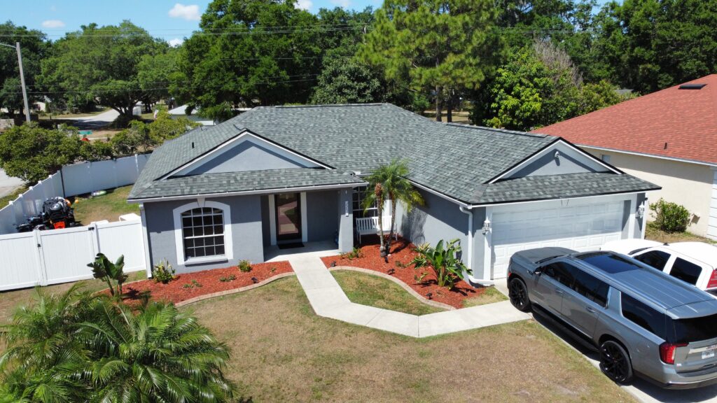 A single-story gray house with a neatly landscaped yard, white trim, and a white fence. Two vehicles are parked in the driveway, and there are trees and greenery surrounding the property.
