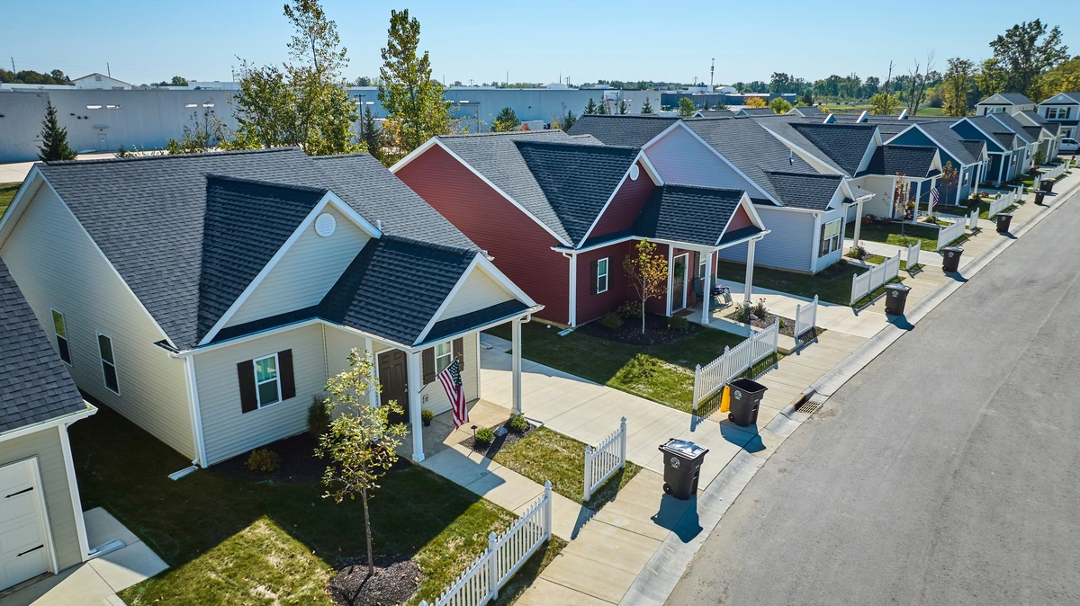 A row of single-story suburban houses with front porches, small lawns, and driveways lines a clean, sunny street. The homes are painted in different colors, and trees dot the yards. A trash bin sits by the curb.