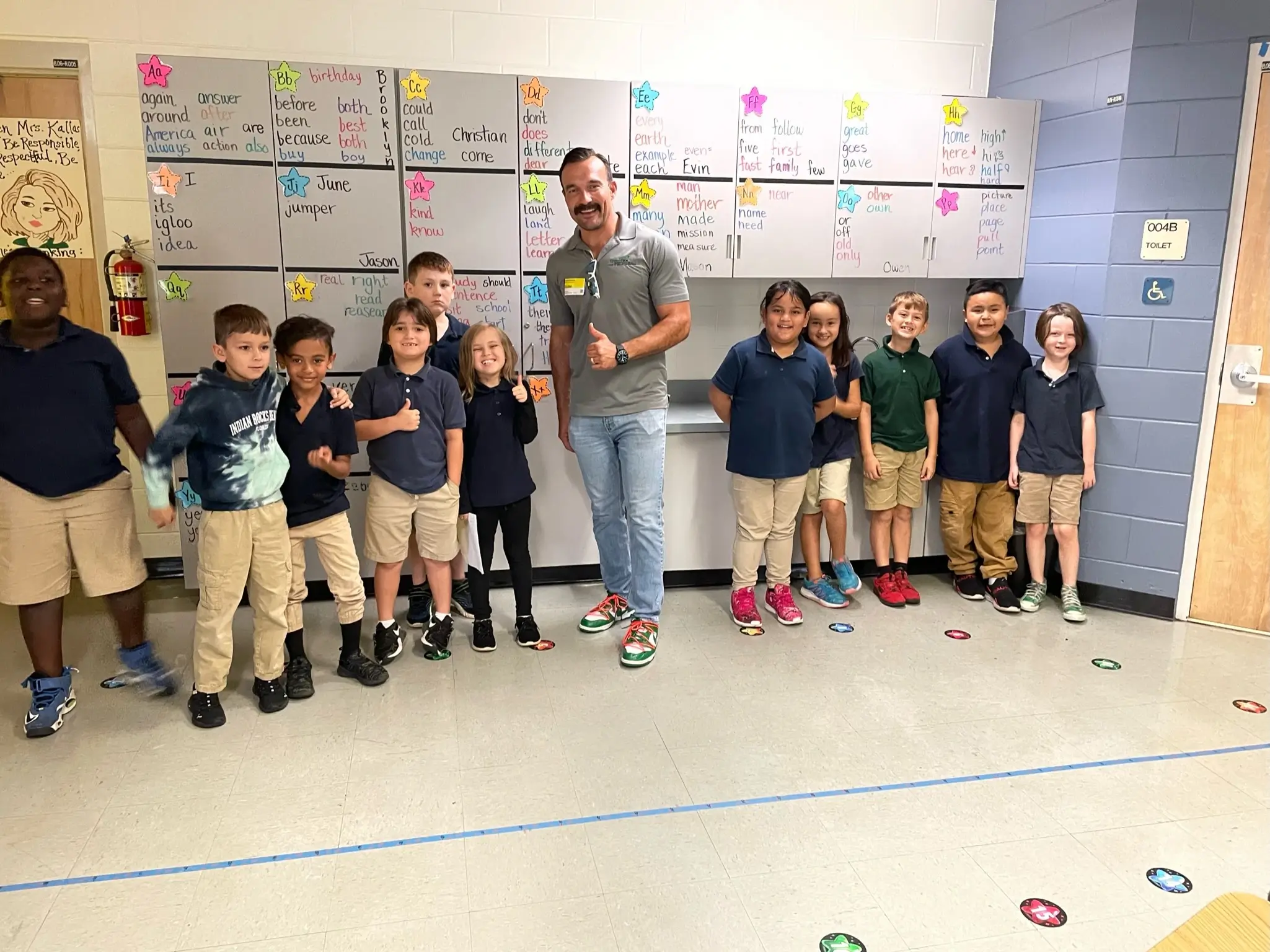 A teacher stands smiling and giving a thumbs up with a group of elementary school students lined up against a classroom wall covered in colorful papers and star-shaped notes.