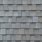 Gray asphalt roof shingles arranged in a staggered, overlapping pattern, creating a textured surface. The shingles have a rough, granular appearance and cover the entire visible area.