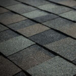 Close-up view of overlapping asphalt roof shingles in shades of gray and brown, arranged in a regular, staggered pattern.