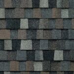 Close-up view of asphalt roof shingles arranged in an overlapping pattern, featuring shades of gray, brown, and black. The textured surface shows slight color variations on each rectangular shingle.