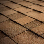 Close-up view of brown asphalt shingles arranged in overlapping rows on a residential roof. The texture of the shingles is visible, showing a mix of light and dark brown tones.