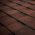 Close-up view of overlapping brown asphalt roof shingles, creating a textured and patterned surface. The lighting highlights the rough granules and the edges of the shingles.