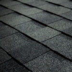 Close-up view of dark asphalt roof shingles arranged in a neat, overlapping pattern. The surface shows a textured, gritty appearance typical of roofing materials.