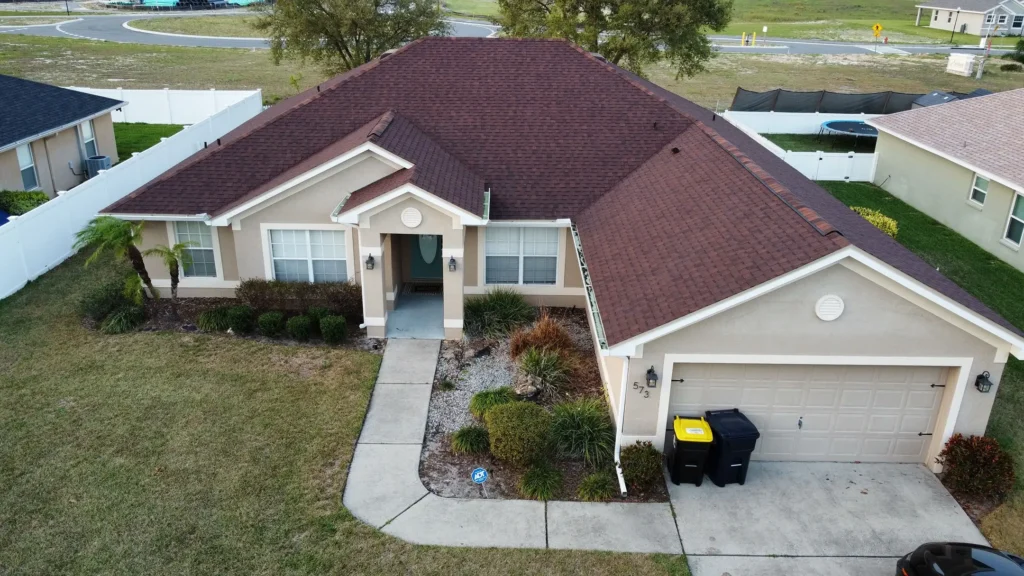 Aerial view of a single-story beige house with a red-brown roof, attached two-car garage, driveway, and well-maintained front yard with shrubs and small trees. Two garbage bins are by the garage.