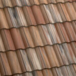 Close-up view of overlapping, wavy terracotta roof tiles with varying shades of brown, red, and tan, creating a textured and weathered appearance.