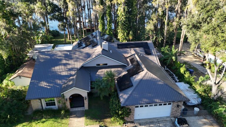 Aerial view of a house with a metal roof, solar panels, and a two-car garage, surrounded by trees and greenery near a body of water. Shadows from the trees cover parts of the roof and yard.