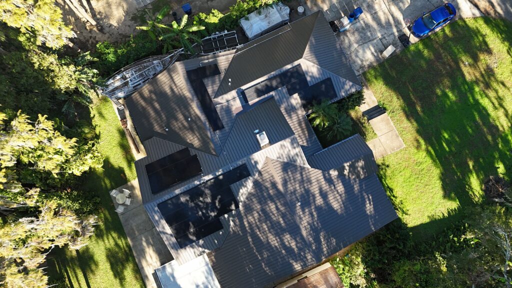 Aerial view of a house with a metal roof and solar panels, surrounded by green lawns, trees, and a driveway with a blue car parked nearby.