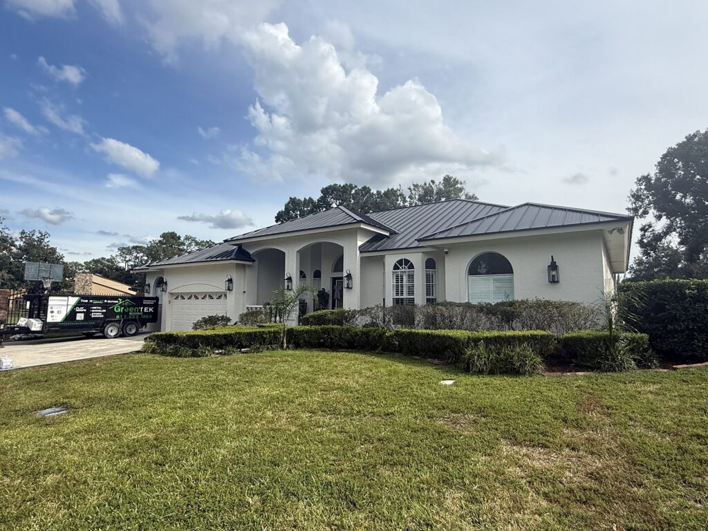 A single-story house with a gray metal roof, white exterior, arched windows, and a large front lawn. A trailer labeled “GUTTER” is parked in the driveway on the left, under a partly cloudy sky.