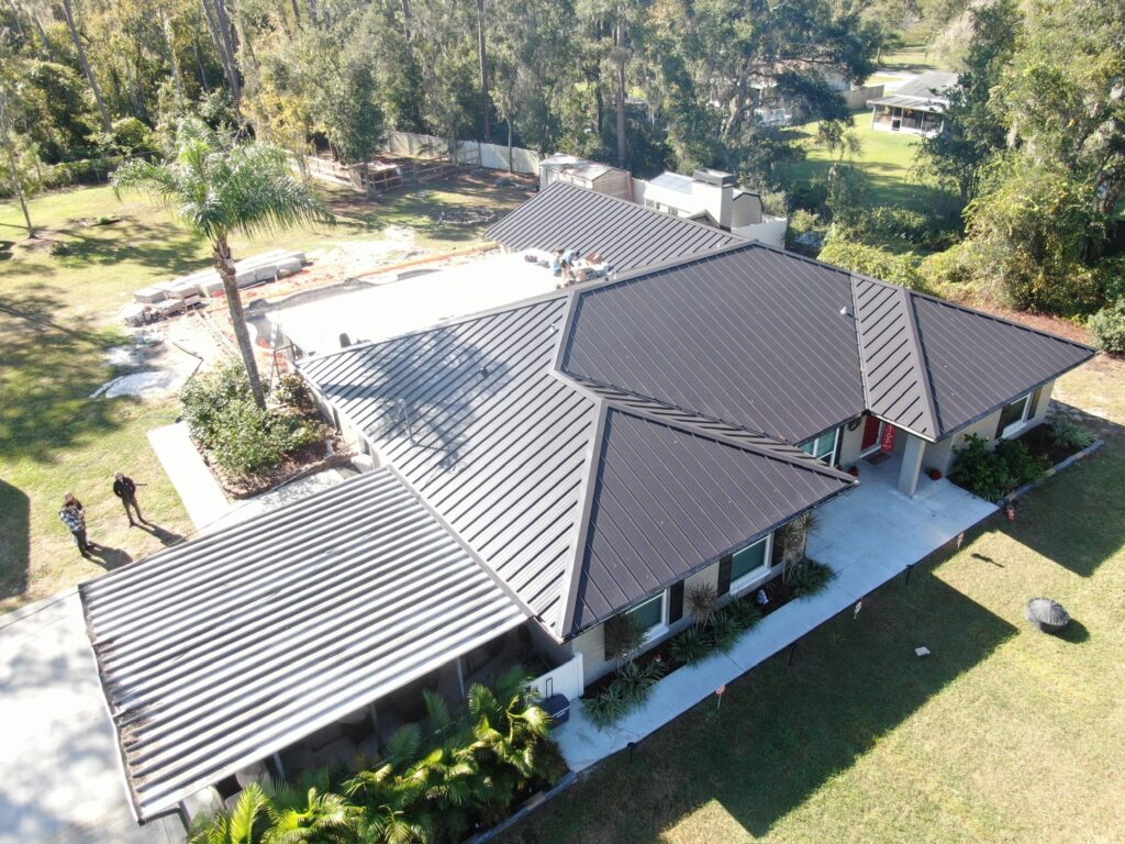 Aerial view of a single-story house with a dark metal roof, surrounded by trees and grass. A driveway, patio, and several people are visible near the house.
