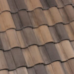Close-up view of overlapping brown and beige clay roof tiles arranged in a wavy, repeating pattern. The tiles have a weathered appearance and varying shades, forming a textured surface.