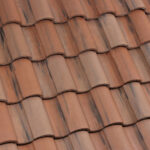 Close-up view of overlapping reddish-brown clay roof tiles with a wavy, textured pattern. The tiles show some darker streaks and natural variation in color, creating a rustic appearance.