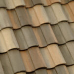 Close-up view of overlapping, weathered clay roof tiles in shades of brown, beige, and tan, arranged in a uniform, repeating pattern.