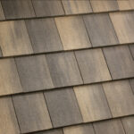A close-up view of overlapping rectangular roof shingles in shades of brown and gray, showing a textured and weathered surface arranged in a diagonal pattern.