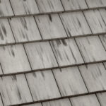 Close-up view of overlapping, rectangular grey roof shingles with visible streaks and weathering marks on the surface.