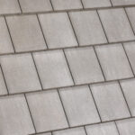Close-up view of a tiled roof with overlapping rectangular gray tiles arranged in a neat, repeating pattern. The tiles have a smooth texture and are laid in horizontal rows.