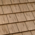 Close-up view of a beige shingle roof with overlapping rectangular tiles arranged in a neat, uniform pattern. The tiles have subtle color variations and streaks, giving them a natural, textured appearance.