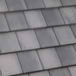 Close-up view of a roof covered with overlapping rectangular gray concrete tiles arranged in neat, uniform rows. The tiles have a slightly textured surface with subtle color variations.