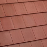 Close-up view of overlapping reddish-brown roof tiles arranged in a uniform, horizontal pattern. The tiles have a smooth, matte texture and are neatly aligned.