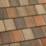 Close-up of a roof with overlapping rectangular shingles in shades of brown, tan, and orange, arranged in a uniform, staggered pattern.