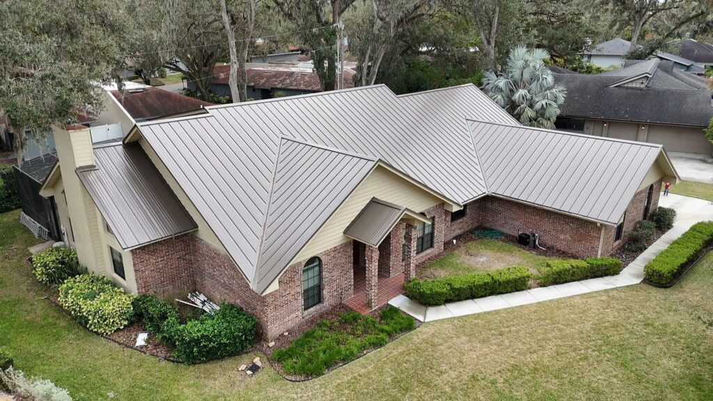 Aerial view of a house with a beige metal roof, brick and tan siding, multiple roof angles, manicured bushes, and a lawn surrounded by trees and neighboring houses.
