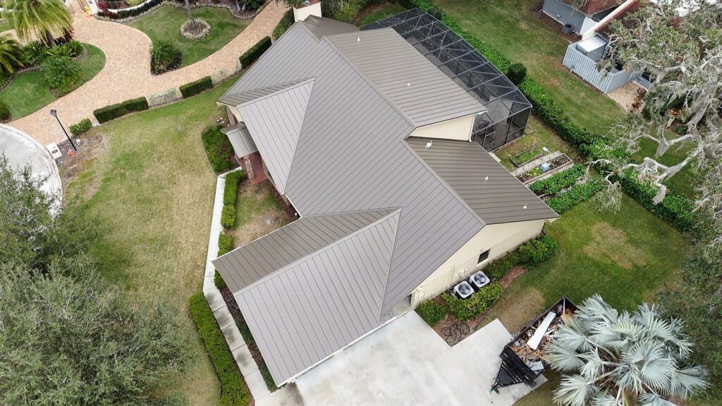 Aerial view of a beige house with a brown metal roof, surrounded by a green lawn, driveway, and garden areas. The backyard features a screened-in patio and various landscaping elements.