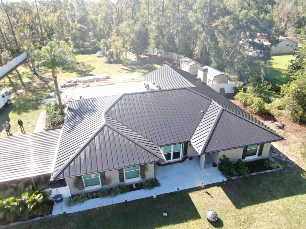Aerial view of a single-story house with a dark metal roof, light-colored exterior walls, and a concrete walkway, surrounded by grass, trees, and a spacious yard.