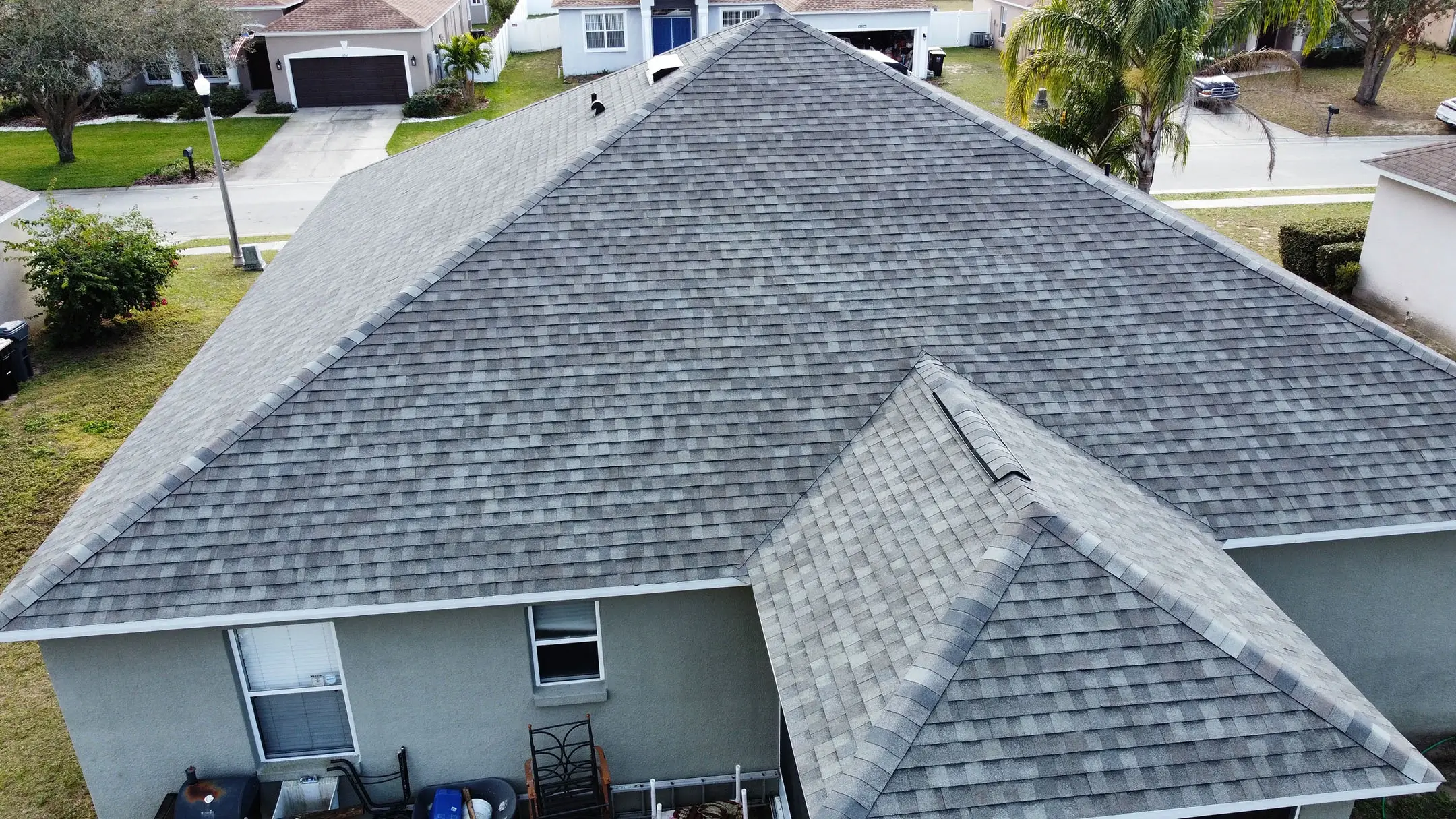 Aerial view of a house with a gray shingle roof, showing the backyard area and nearby residential homes, with palm trees and green lawns visible in the neighborhood.