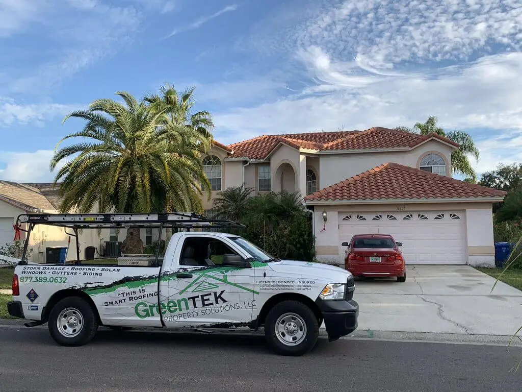 A GreenTek pickup truck with a ladder on top is parked in front of a beige two-story house with a red-tiled roof, palm trees, and a red car in the driveway on a sunny day.